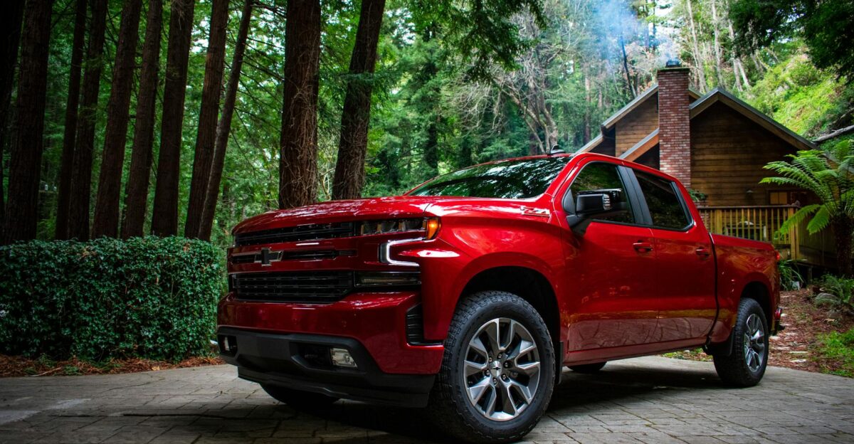 A red Chevrolet Silverado 1500 parked in a rustic driveway surrounded by lush forest and cottage